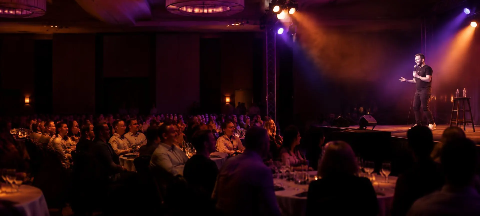 Corporate ballroom with round tables and a truss-lit stage, audience laughing